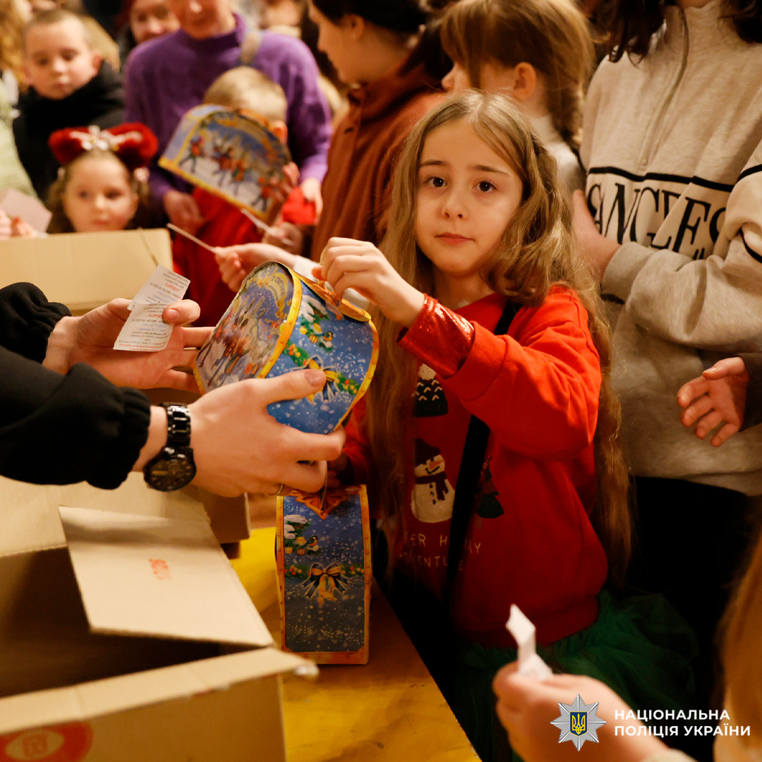 У приміщенні під час святкового заходу працівник Національної поліції України передає дитині яскраву подарункову коробку з новорічними ілюстраціями. На задньому плані видно інших дітей та дорослих, а також коробки з подарунками.
