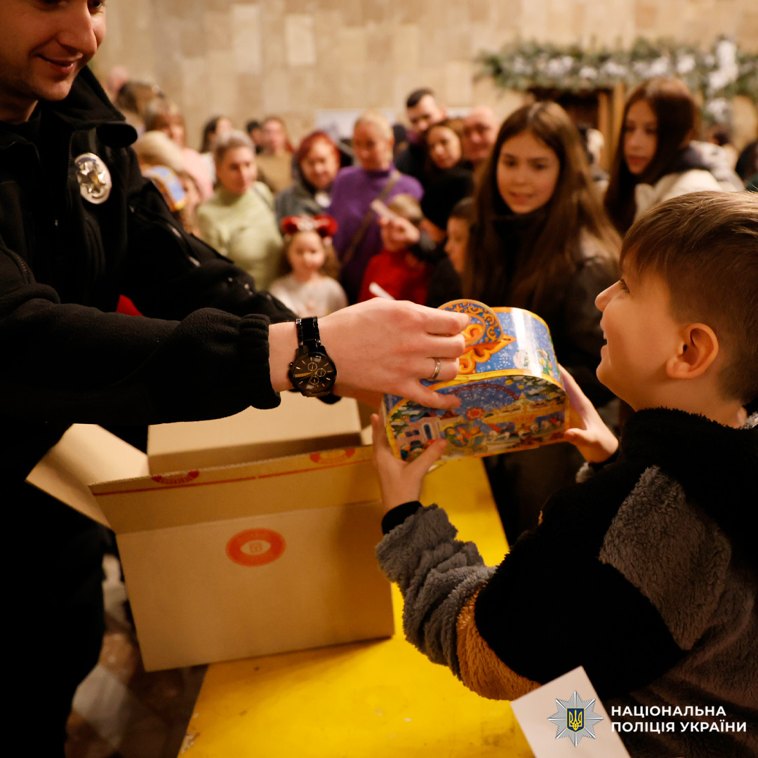 У приміщенні під час святкового заходу працівник Національної поліції України передає дитині яскраву подарункову коробку з новорічними ілюстраціями. На задньому плані видно інших дітей та дорослих, а також коробки з подарунками.