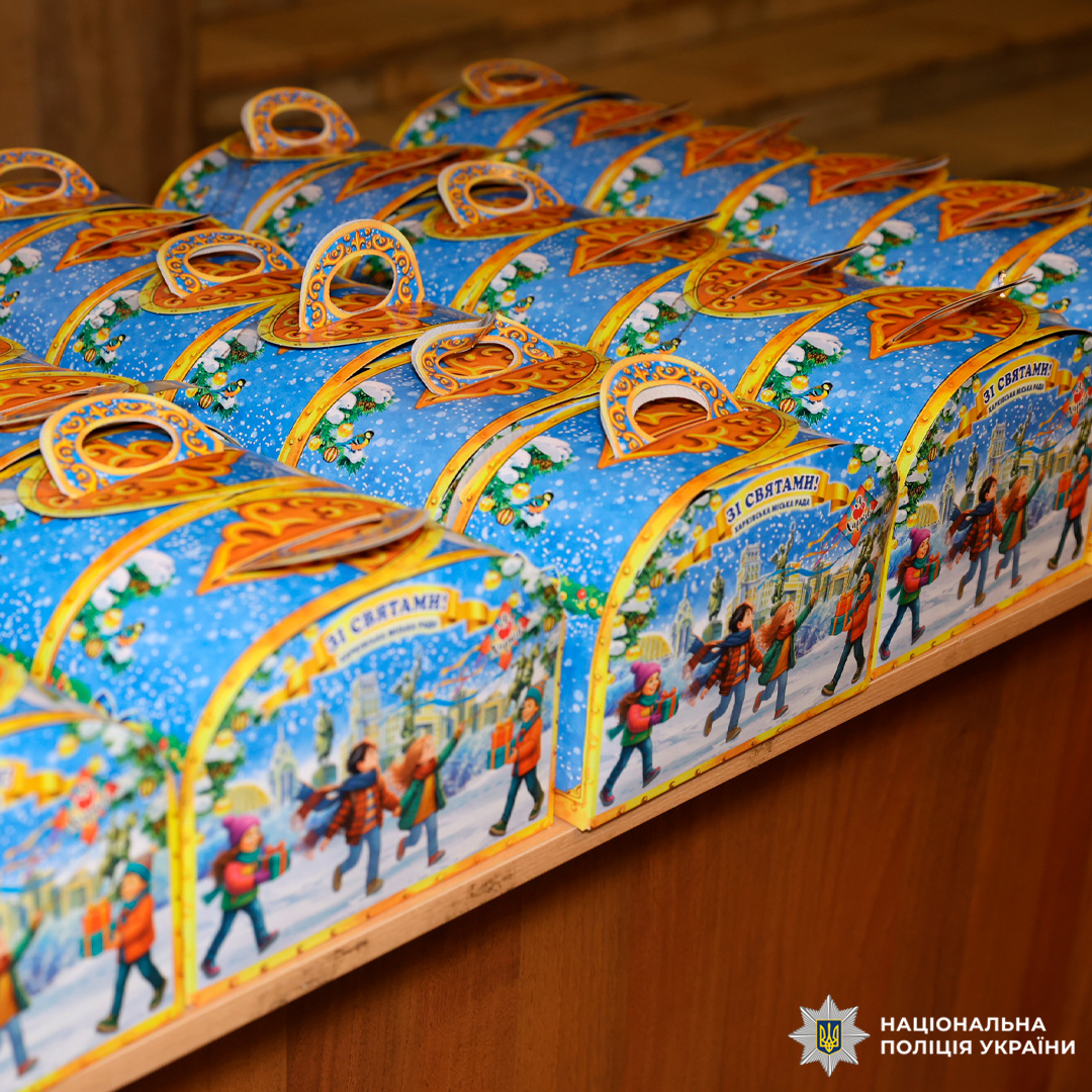 На дерев’яній поверхні акуратно розташовані святкові коробки синього кольору з ілюстраціями зимового міста та написом 'Зі святами!'. Коробки мають ручки для перенесення.