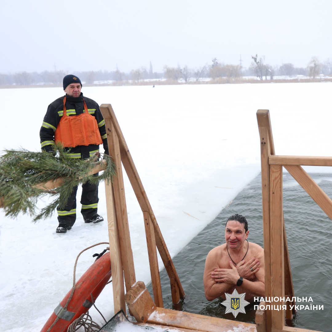 Чоловік занурюється в ополонку, поруч стоїть рятувальник у зимовій формі.