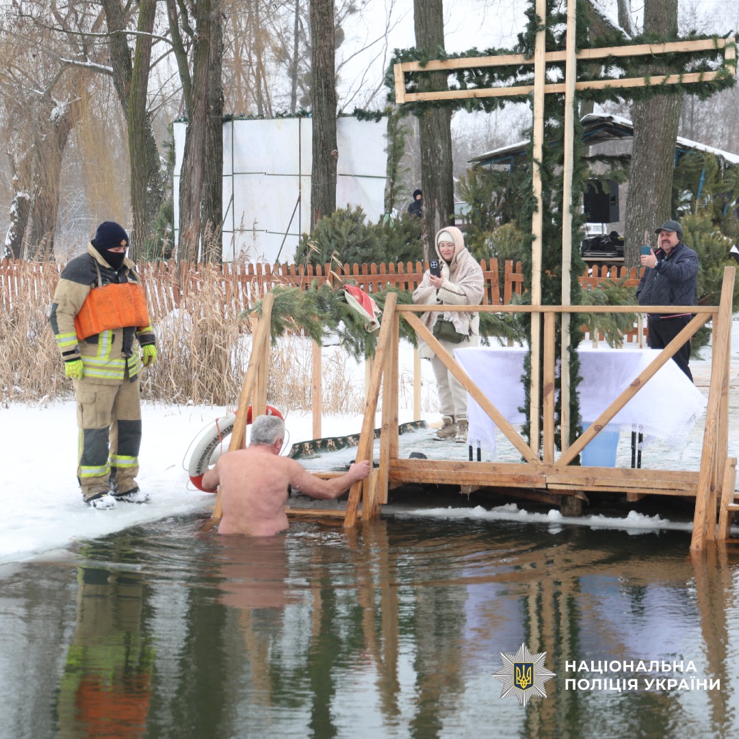 Чоловік занурюється в ополонку, поруч стоїть рятувальник у зимовій формі.