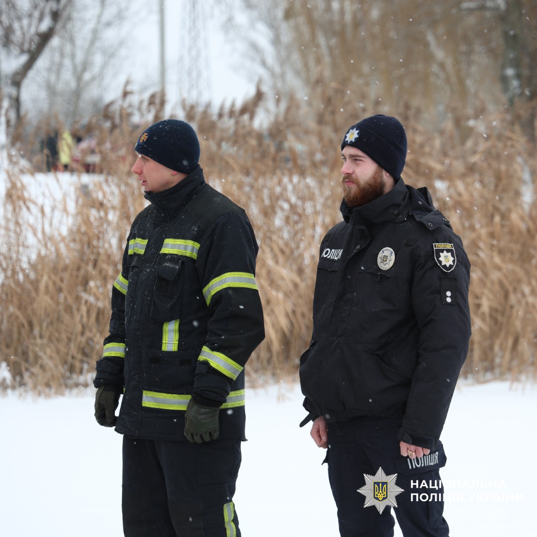 Поліцейський та рятувальник у зимовій формі стоять на засніженій місцевості біля очерету.