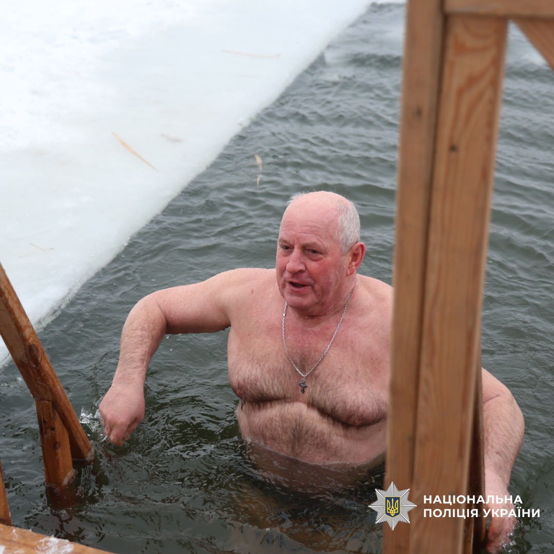 Літній чоловік занурюється в ополонку, тримаючись за дерев’яну конструкцію.