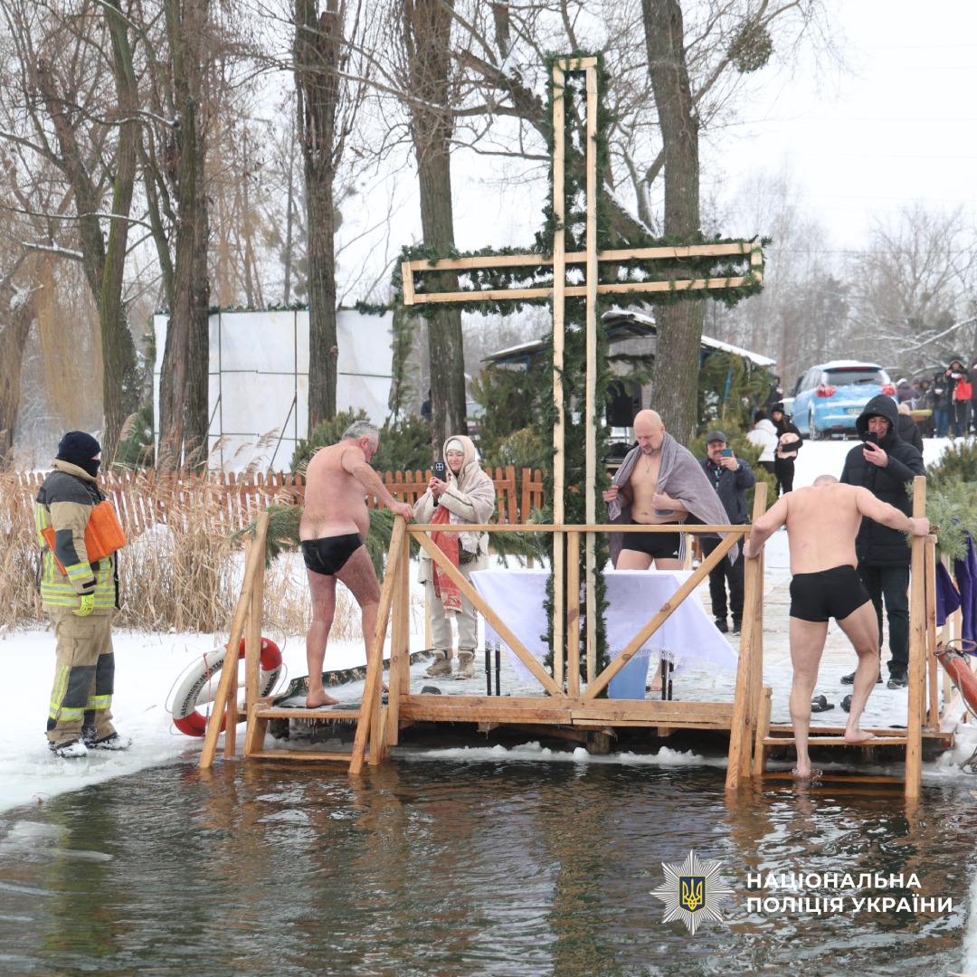 Група людей бере участь у зимовому купанні біля прикрашеного хрестом помосту, присутні рятувальники та поліція.