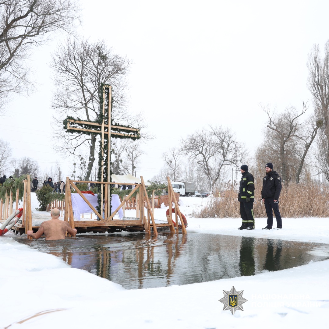 Група людей бере участь у зимовому купанні біля прикрашеного хрестом помосту, присутні рятувальники та поліція.