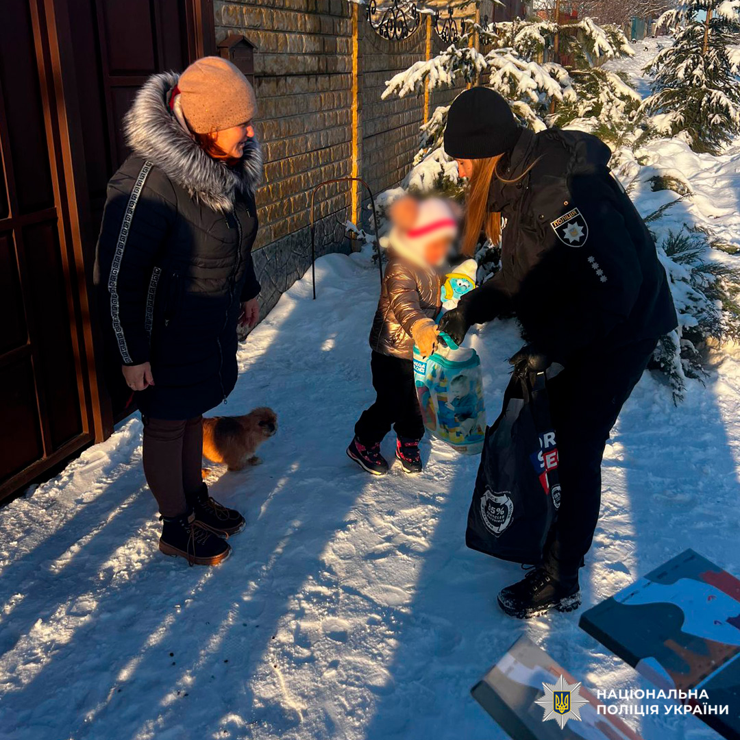 Поліцейський передає дитині контейнер із подарунками, поруч стоїть дорослий та маленька собака.