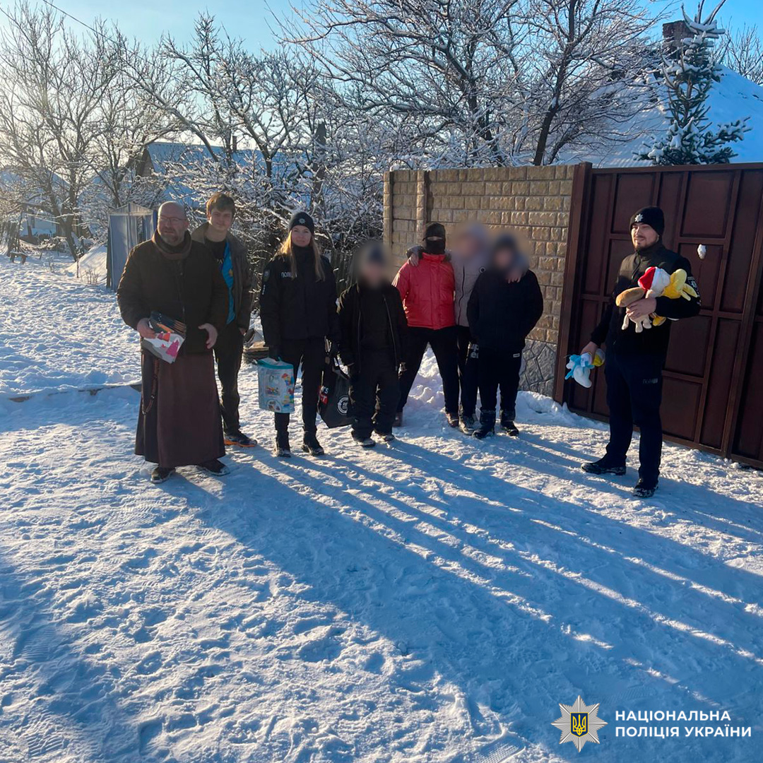 Група людей із подарунками та іграшками стоїть на засніженій території, серед них поліцейський і священнослужитель.