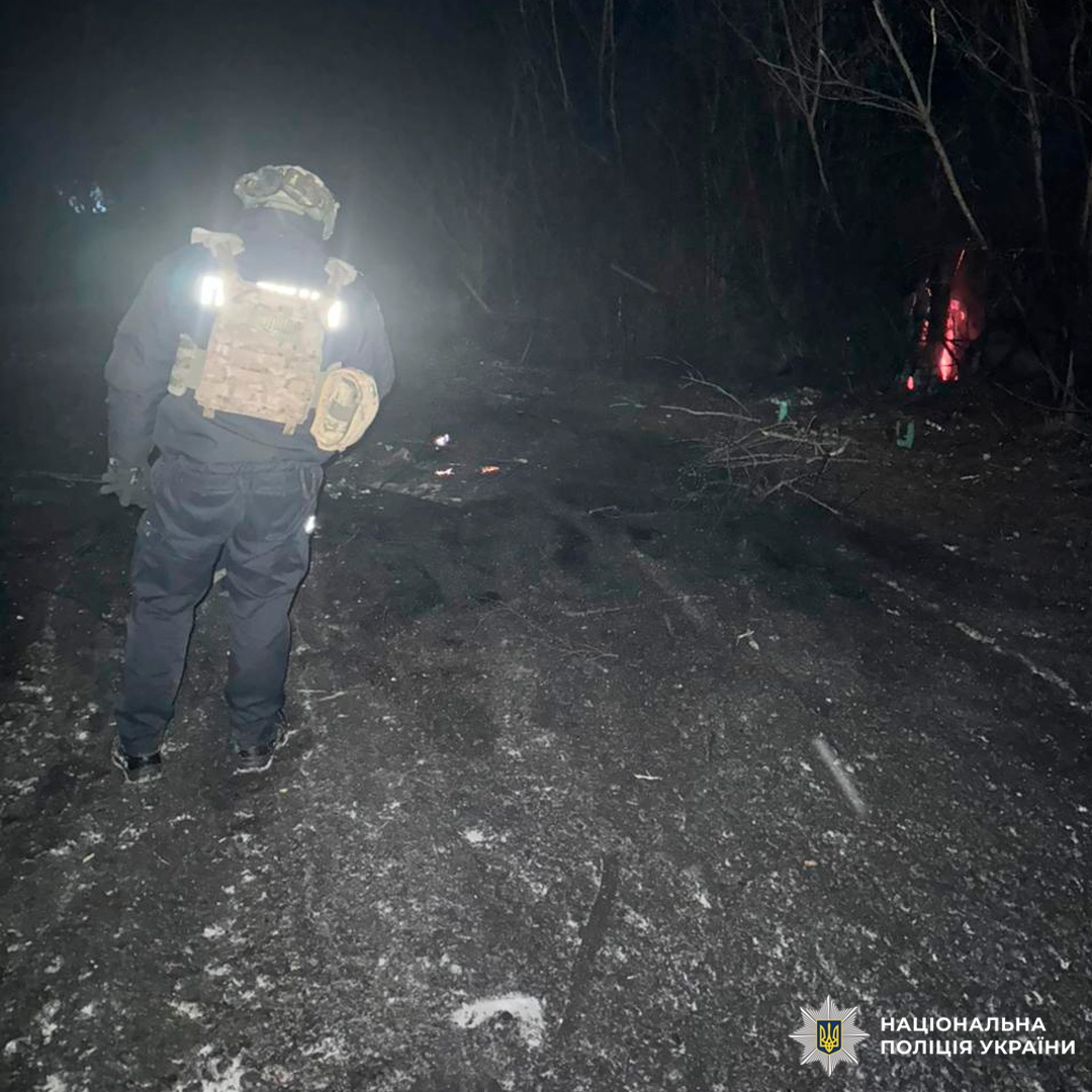 Поліцейський оглядає місце події, освітлюючи тріщини на землі ліхтарем у нічний час.
