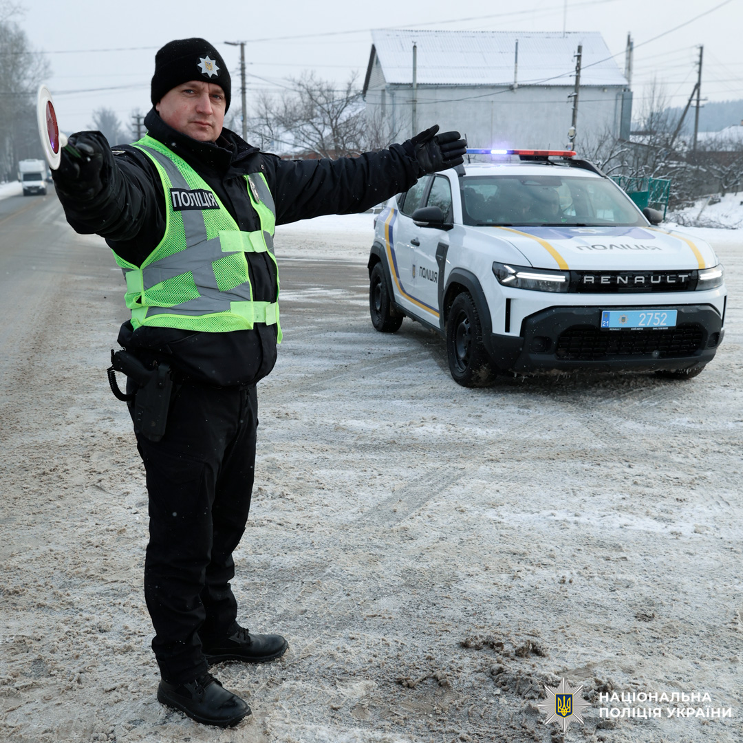 Поліцейський регулює рух транспорту на засніженій дорозі поруч із службовим автомобілем з увімкненими проблисковими маячками.