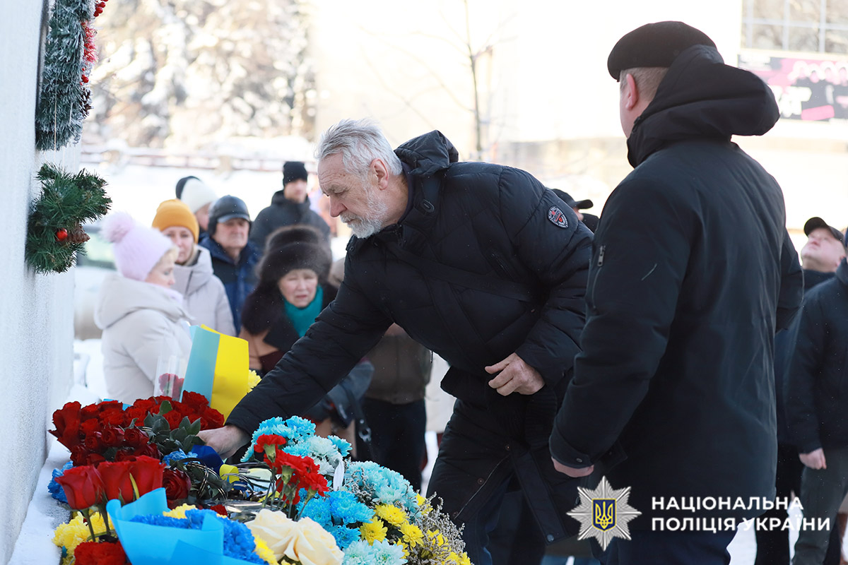 На фото люди кладуть жовто-блакитні квіти до меморіальної дошки