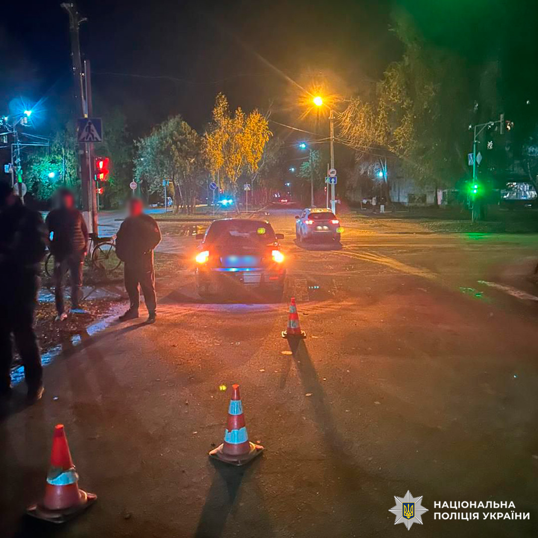 Поліцейські задокументували дорожньо-транспортну пригоду на перехресті вночі. Автомобіль із увімкненими аварійними сигналами оточений конусами, поруч перебувають кілька людей.