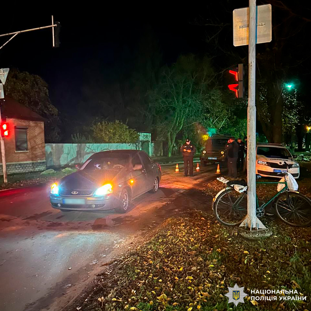 Поліцейські задокументували дорожньо-транспортну пригоду на перехресті вночі. У центрі дороги зупинився легковий автомобіль із увімкненими фарами та аварійними сигналами, навколо встановлені конуси. Поруч працюють правоохоронці, неподалік стоїть службовий автомобіль.