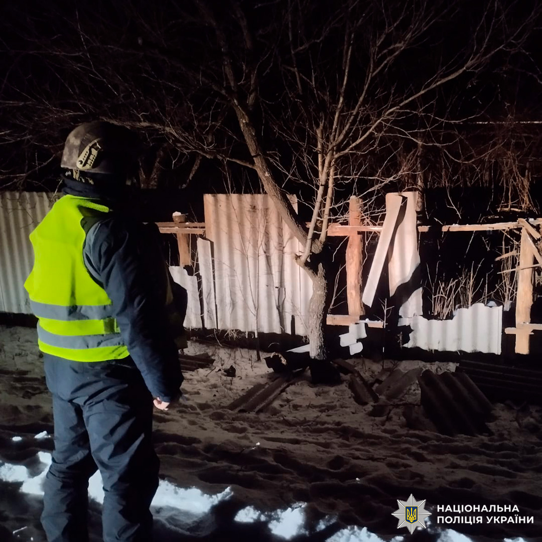 Працівник поліції у світловідбивному жилеті та шоломі стоїть біля пошкодженого металевого паркану вночі. На землі лежить сніг, за парканом видно дерева без листя.