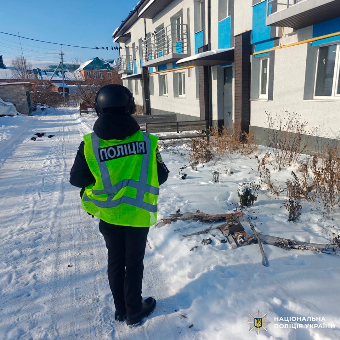 Працівник поліції у світловідбивному жилеті з написом «ПОЛІЦІЯ» стоїть на засніженій доріжці біля багатоповерхового житлового будинку. На землі видно уламки та суху рослинність.