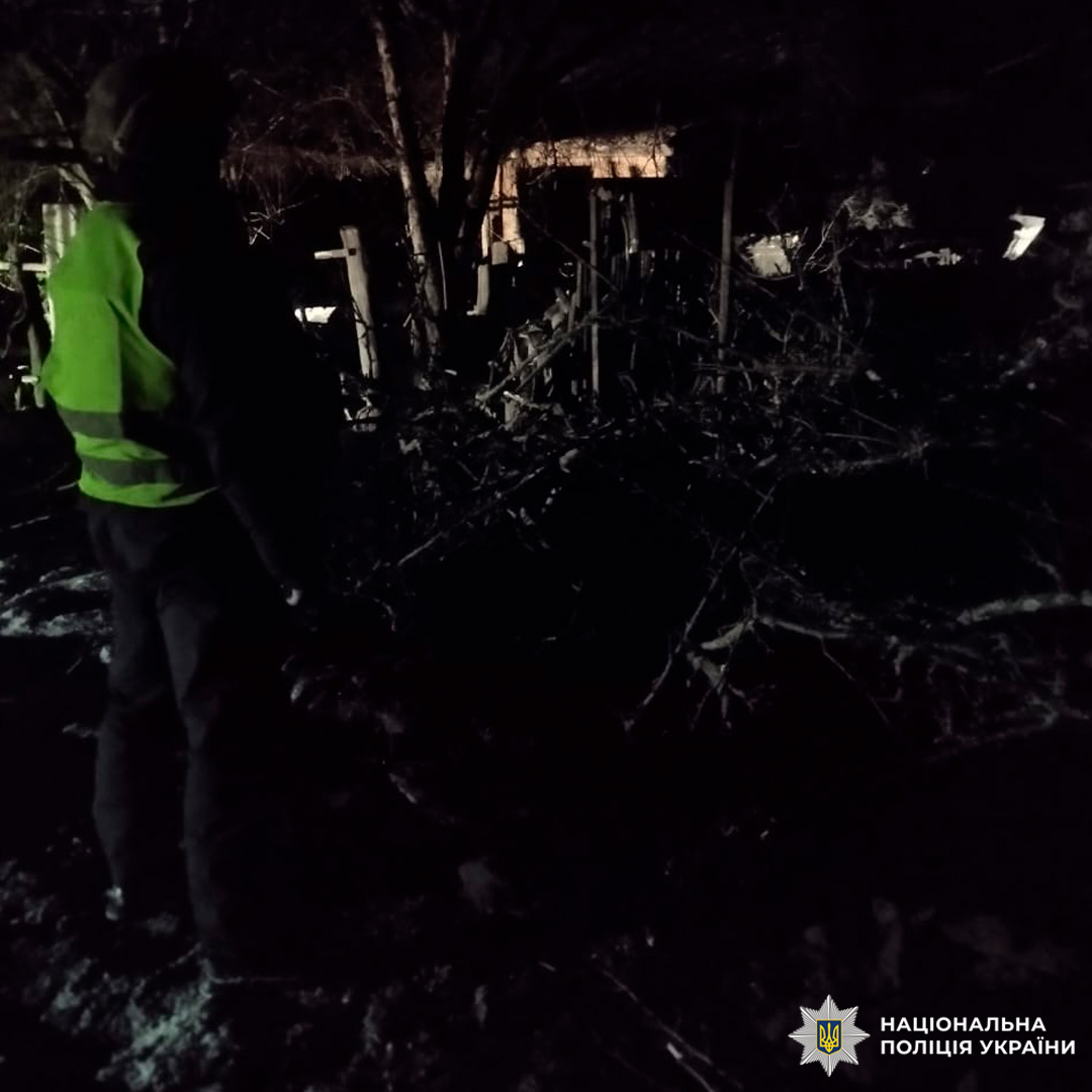 Працівник поліції у світловідбивному жилеті стоїть у темній місцевості серед дерев та огорожі. На землі лежить сніг або іній.