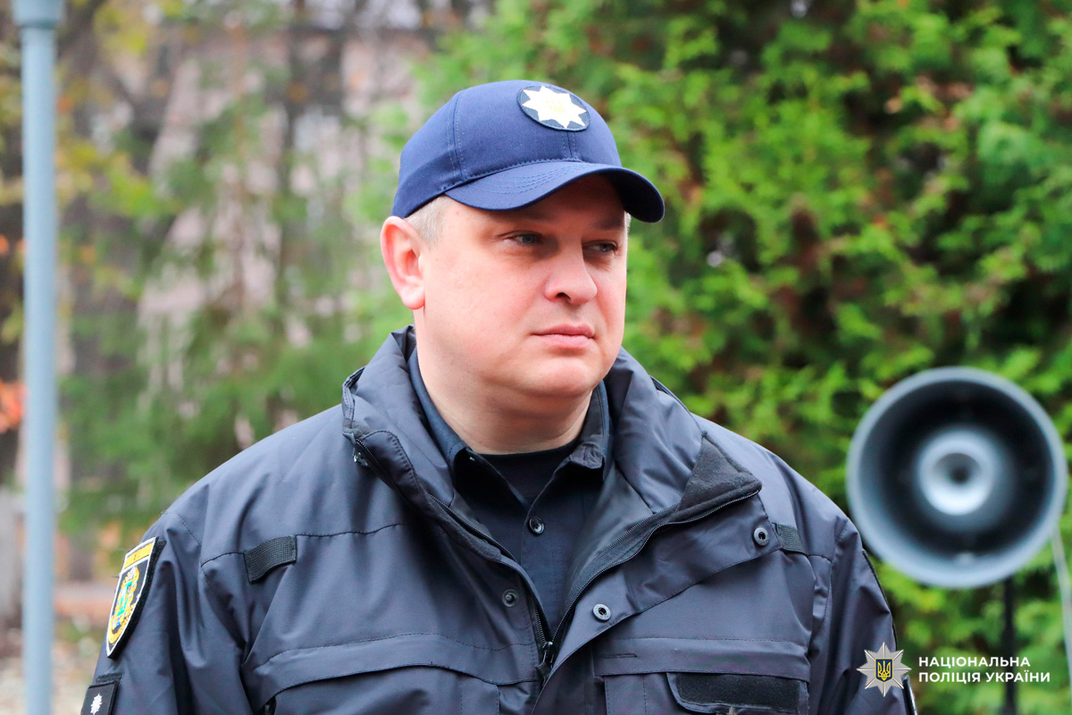 Поліцейський у формі Національної поліції України стоїть на тлі зелених насаджень.