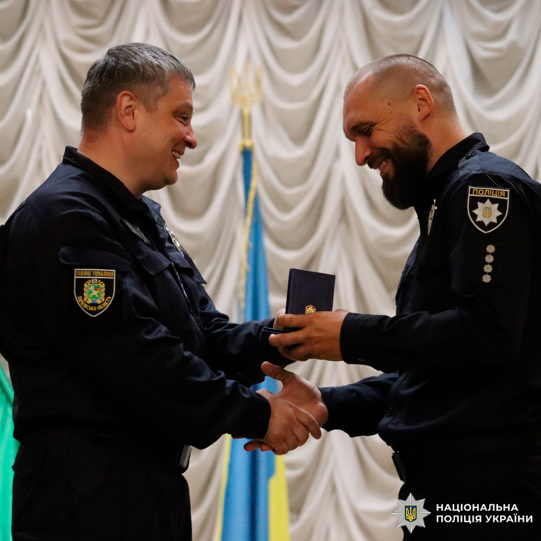 Двоє співробітників Національної поліції України стоять на сцені перед прапором України. Один офіцер передає іншому нагороду у синьому футлярі, супроводжуючи це рукостисканням. Обидва усміхаються.
