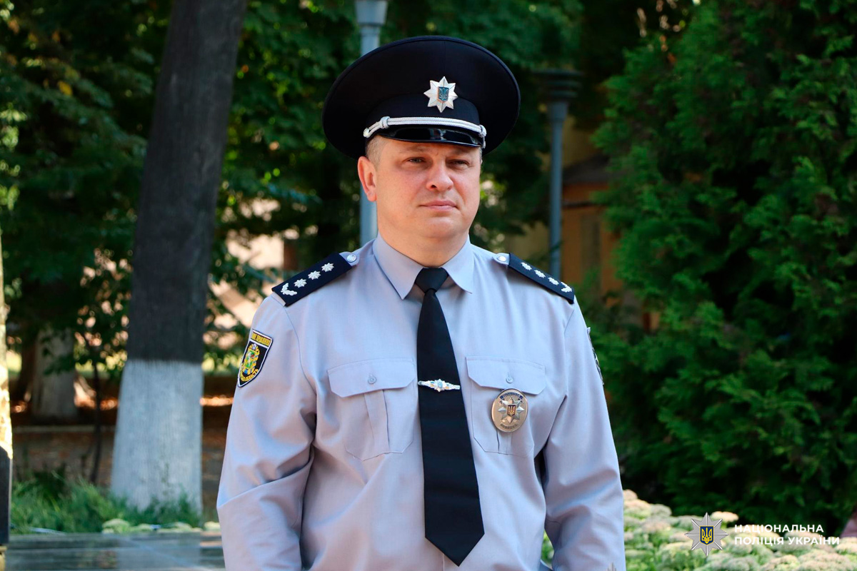 Співробітник Національної поліції України у світло-блакитній сорочці з еполетами та срібним кашкетом стоїть на відкритому повітрі перед зеленим фоном. На формі видно нагрудний знак, що підкреслює офіційний характер зображення.