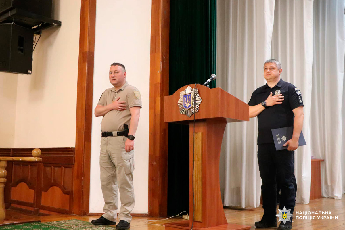 Двоє чоловіків у формі стоять на сцені з руками на серці біля трибуни з гербом України.