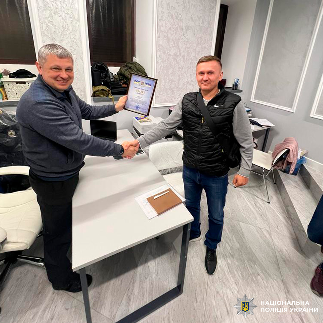 У сучасному офісному приміщенні співробітник Національної поліції України та цивільний обмінюються рукостисканням. Один передає іншому рамку з грамотою, що свідчить про офіційне вручення відзнаки.