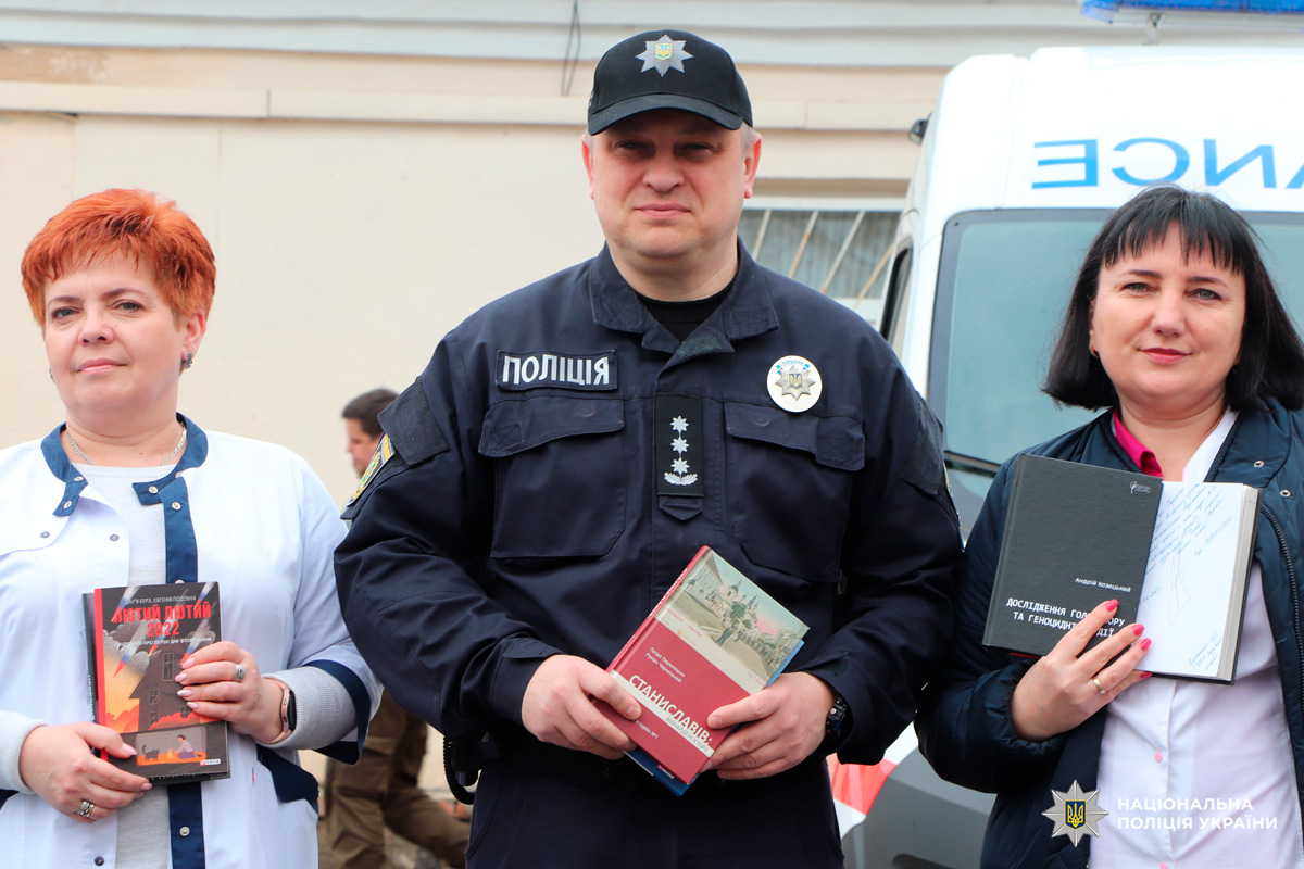 Поліцейський та двоє цивільних тримають книги на тлі автомобіля швидкої допомоги.