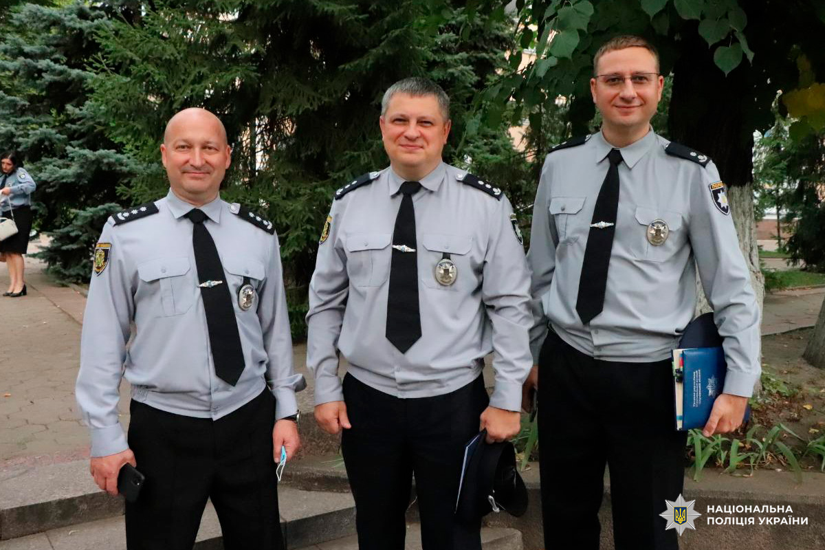 Троє офіцерів Національної поліції України у світло-сірих сорочках стоять на відкритому повітрі.
