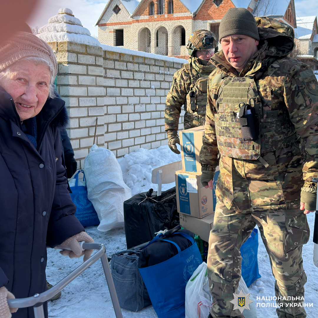Поліцейські у камуфляжній формі допомагають роздавати гуманітарну допомогу літній людині з ходунками.