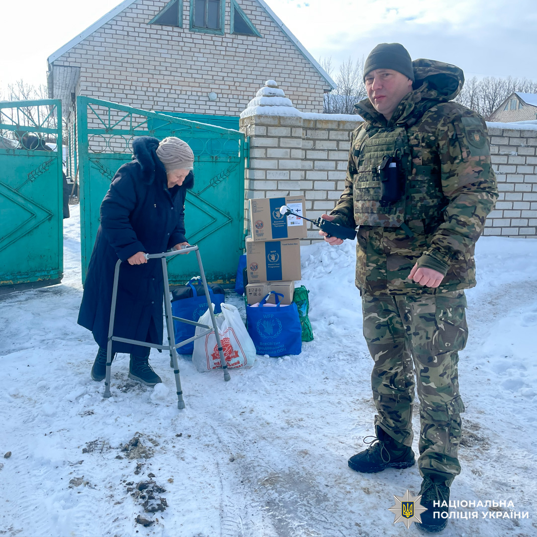 Поліцейські у камуфляжній формі допомагають роздавати гуманітарну допомогу літній людині з ходунками.