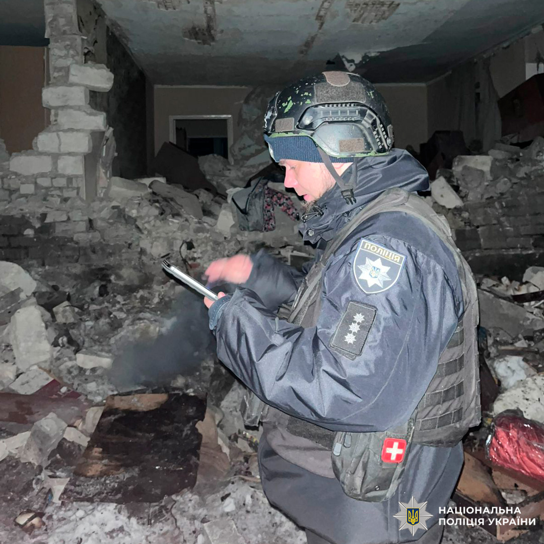 Поліцейський у тактичному спорядженні стоїть серед уламків у задимленому середовищі, дивиться на мобільний пристрій.