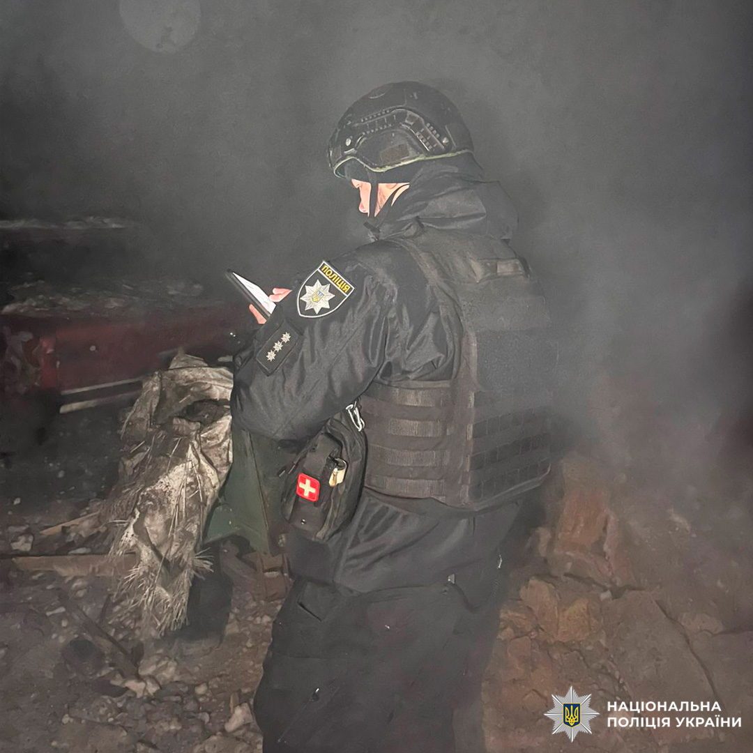 Поліцейський у тактичному спорядженні стоїть серед уламків у задимленому середовищі, дивиться на мобільний пристрій.