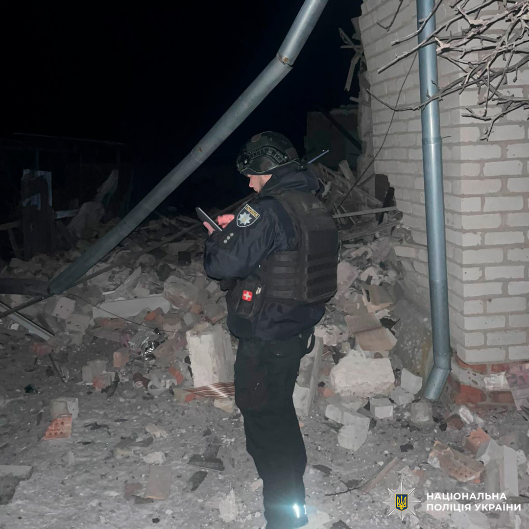 Поліцейський у тактичному спорядженні стоїть серед уламків у задимленому середовищі, дивиться на мобільний пристрій.