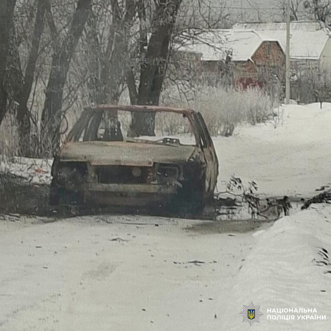 Згорілий автомобіль стоїть на засніженій сільській дорозі, навколо дерева та будинки з білими дахами.