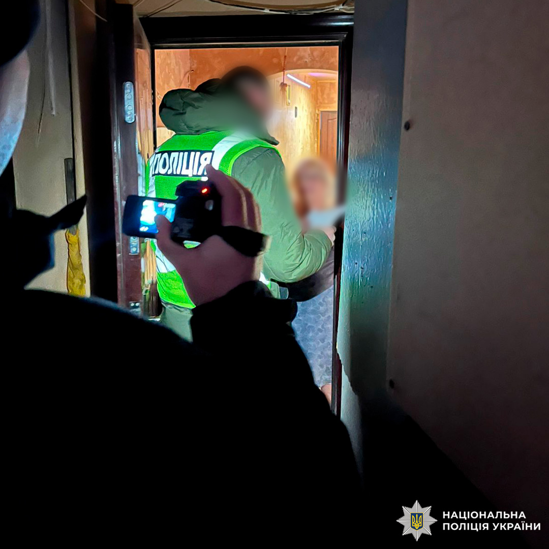 Поліцейський у жилеті «Поліція» взаємодіє з особою у коридорі житлового будинку, поруч оператор із камерою.