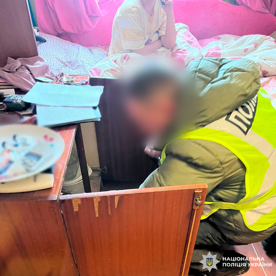 Поліцейський у жилеті «Поліція» оглядає вміст шафи, поруч у кімнаті сидить людина на ліжку.