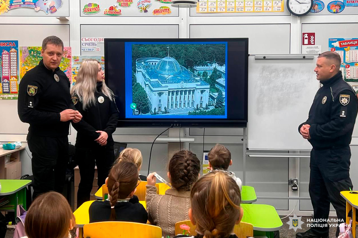 Троє поліцейських у формі Національної поліції України проводять заняття для дітей у класі. На екрані видно аерофотознімок будівлі з куполом. Діти сидять за кольоровими партами та уважно слухають.