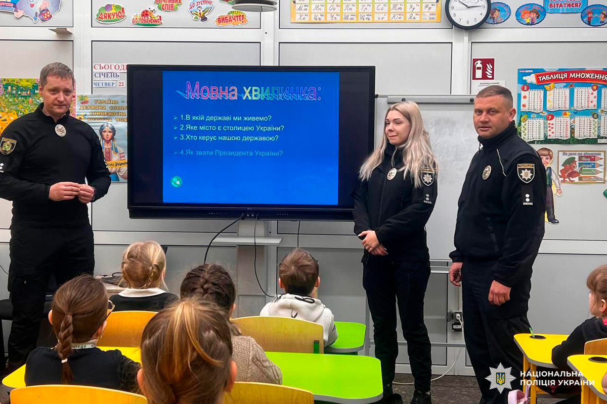 Троє поліцейських Національної поліції України проводять навчальне заняття для школярів у класі. На екрані демонструється слайд «Мовна хвилинка» з чотирма запитаннями про державу та її керівництво. Діти сидять за кольоровими партами й уважно слухають.