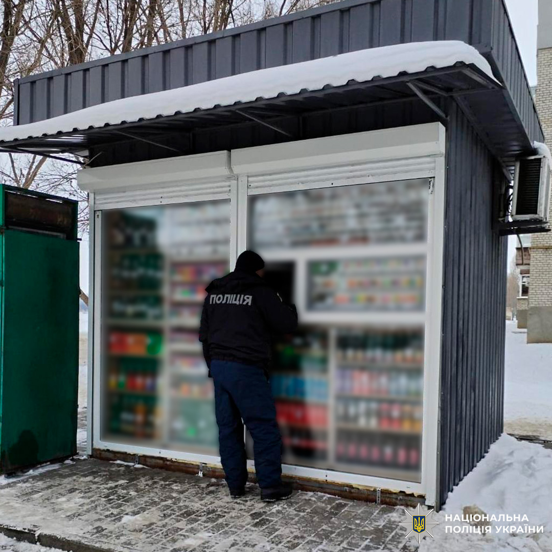 У зимових умовах на засніженій вулиці поліцейський у темній формі стоїть перед кіоском із прозорими вітринами. Усередині видно ряди напоїв та полиці з пачками сигарет.