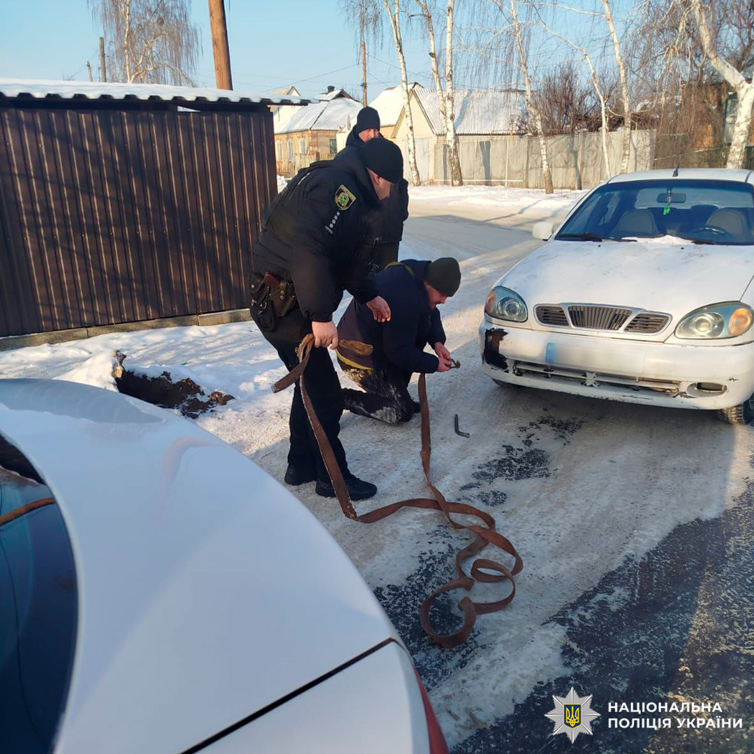 У зимових умовах двоє чоловіків, серед яких поліцейський у формі, прикріплюють буксирувальний трос до білого автомобіля з пошкодженим переднім бампером. Один із них працює навколішки, інший допомагає поруч. Третій чоловік стоїть позаду, спостерігаючи. Навколо видно сніг, дерев’яний сарай та будинки з засніженими дахами.