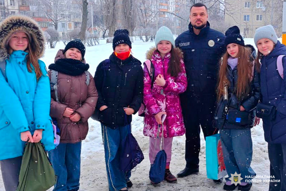 Поліцейський у формі стоїть поруч із групою учнів на засніженій вулиці житлового району.