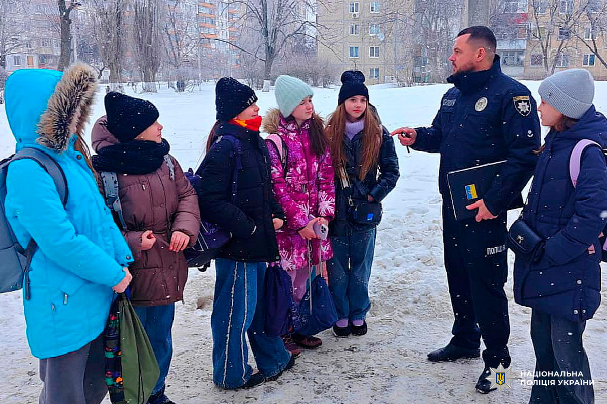 Поліцейський у формі стоїть поруч із групою учнів на засніженій вулиці житлового району.