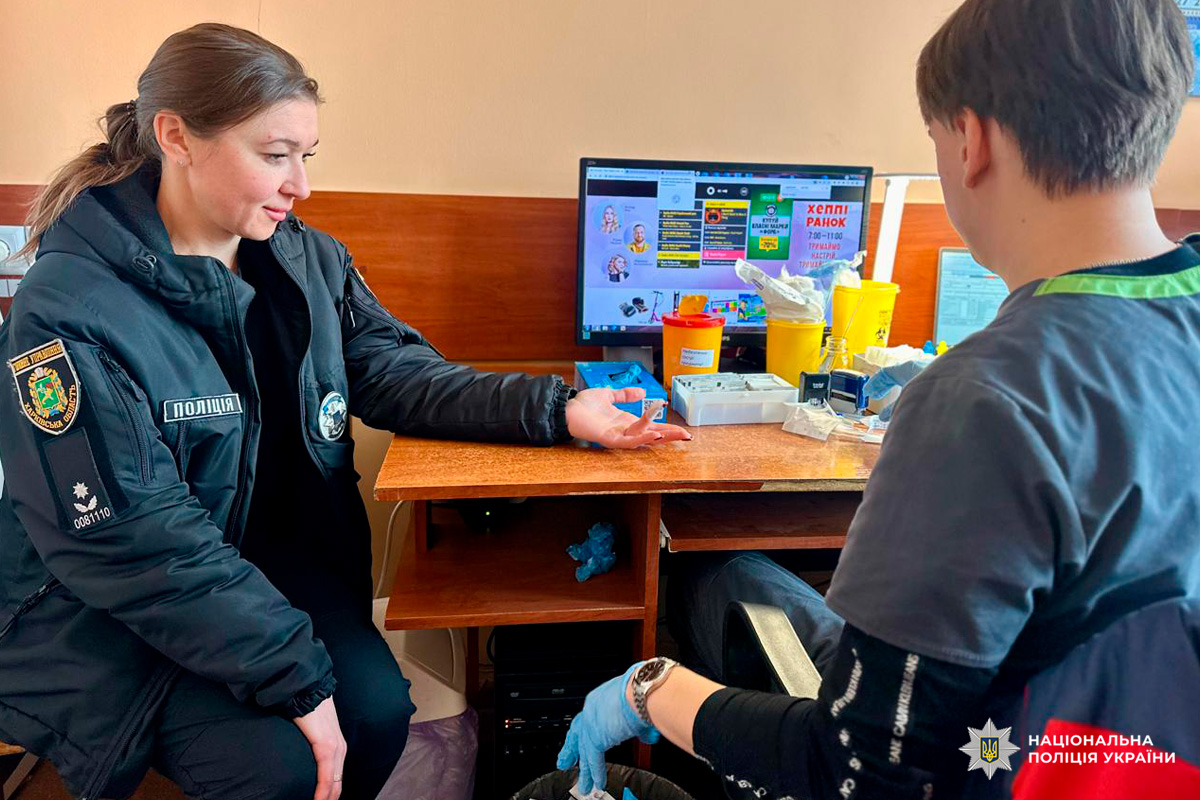 У приміщенні поліцейська у формі сидить за столом із витягнутою рукою, медичний працівник у рукавичках готується взяти зразок. На столі видно медичні матеріали, у фоні - комп’ютерний монітор.