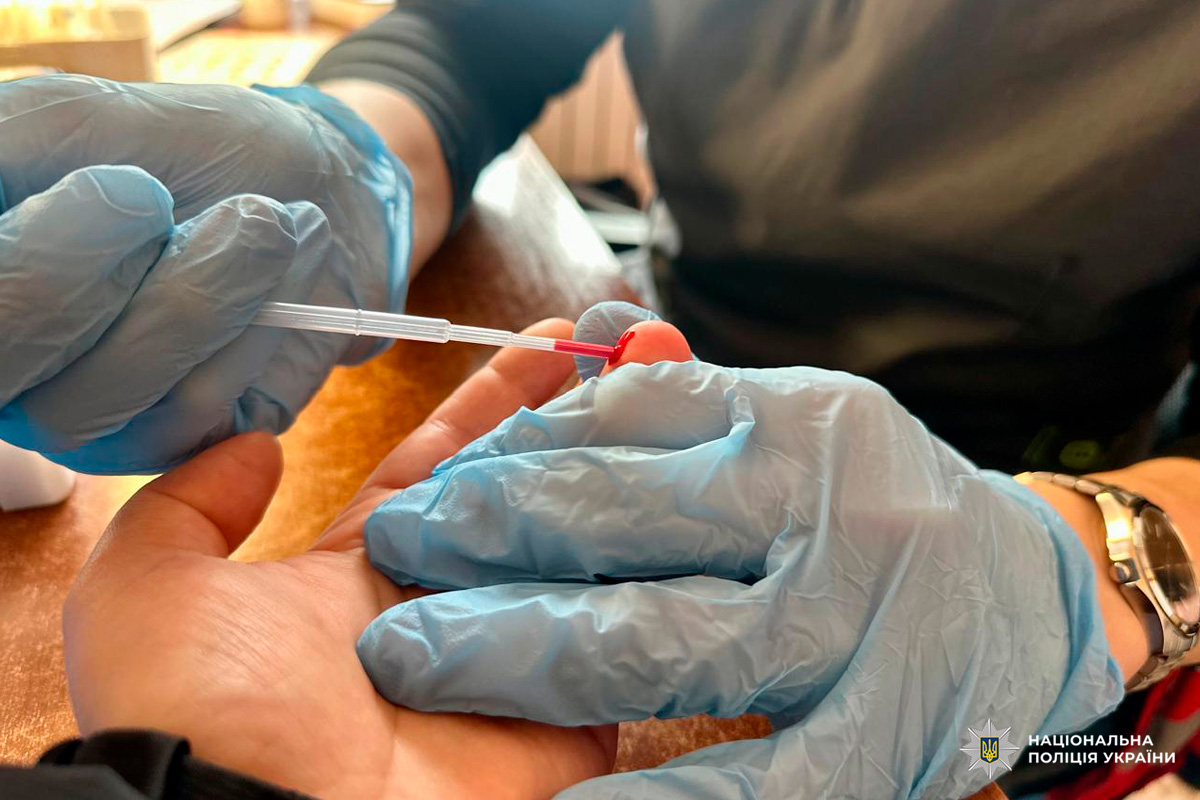 У приміщенні медичний працівник у синіх рукавичках використовує піпетку для забору краплі крові з пальця.