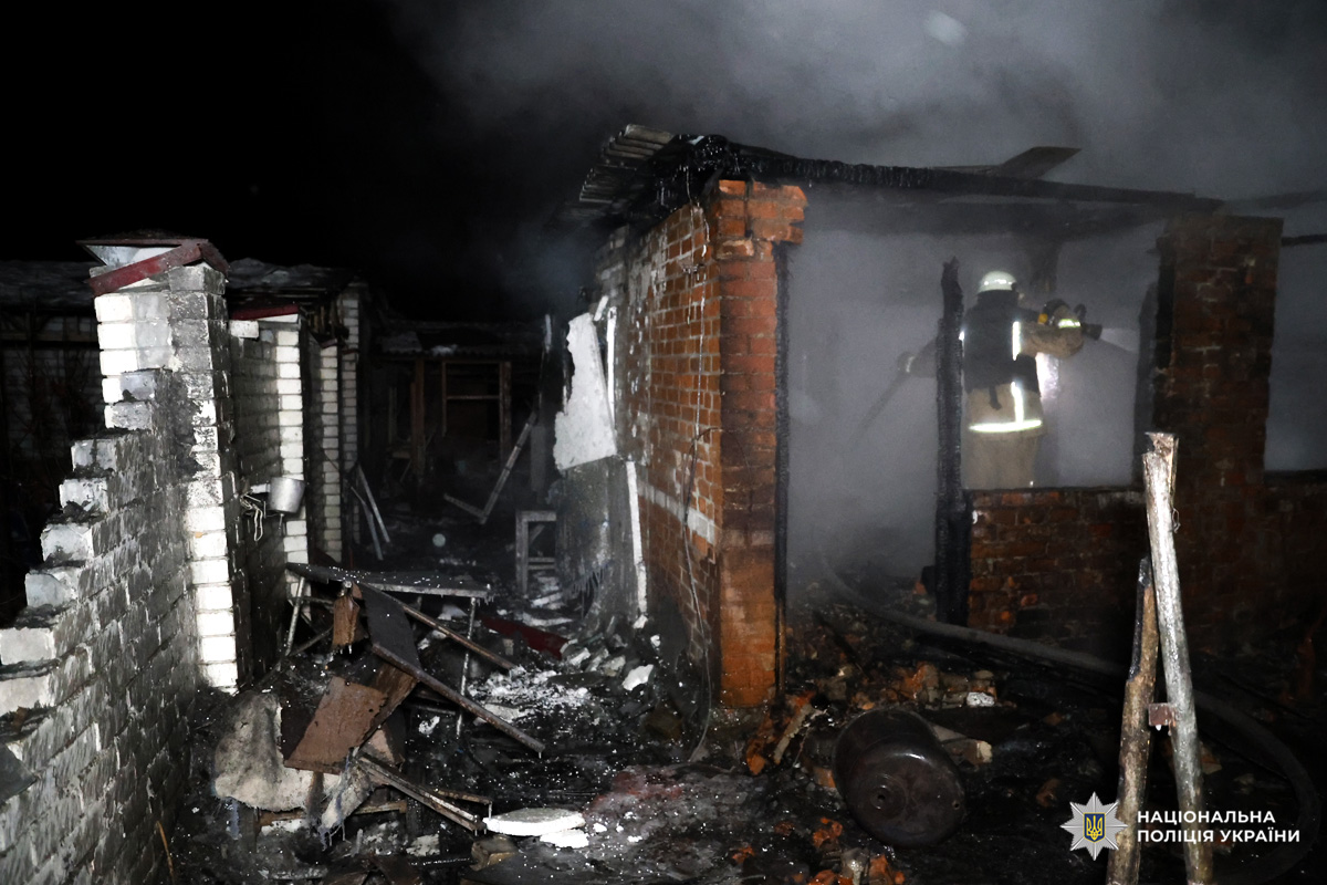 Цегляна будівля з обгорілими стінами та уламками, пожежник гасить залишки вогню.