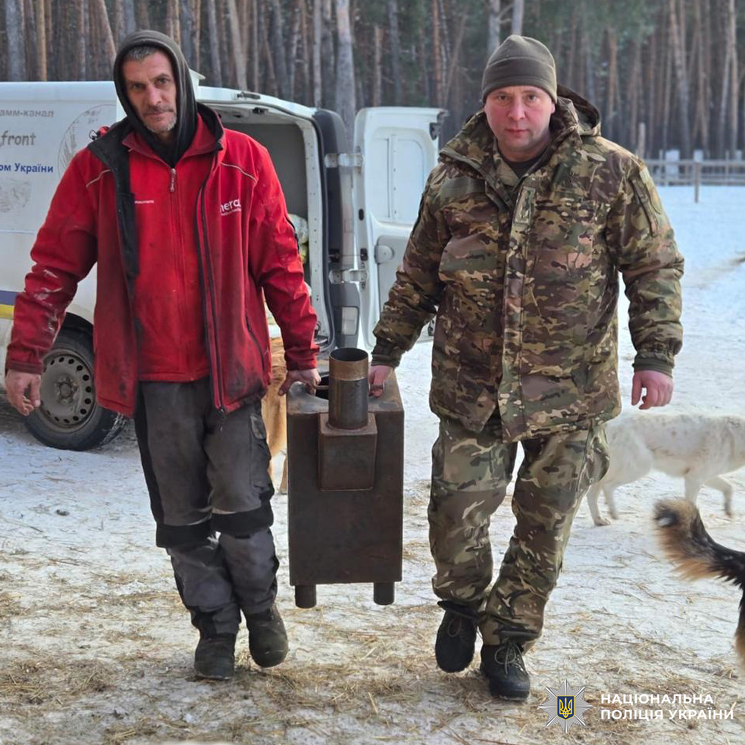 Двоє чоловіків несуть буржуйку взимку, поруч авто та собаки.