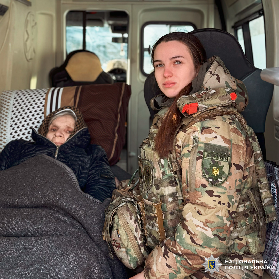 Поліцейський у камуфляжі поруч із літньою людиною в автомобілі швидкої допомоги.