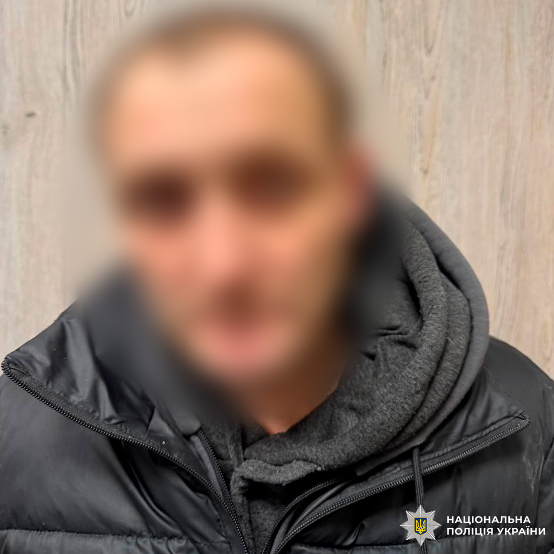 Чоловік у чорній куртці позує на фоні дерев’яної стіни.