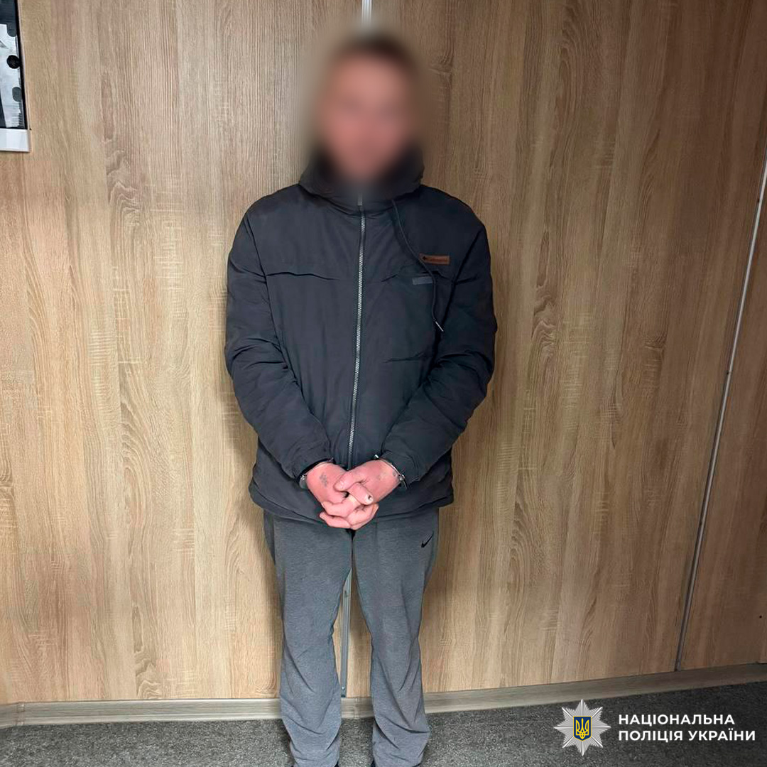 Чоловік у чорній куртці позує на фоні дерев’яної стіни.