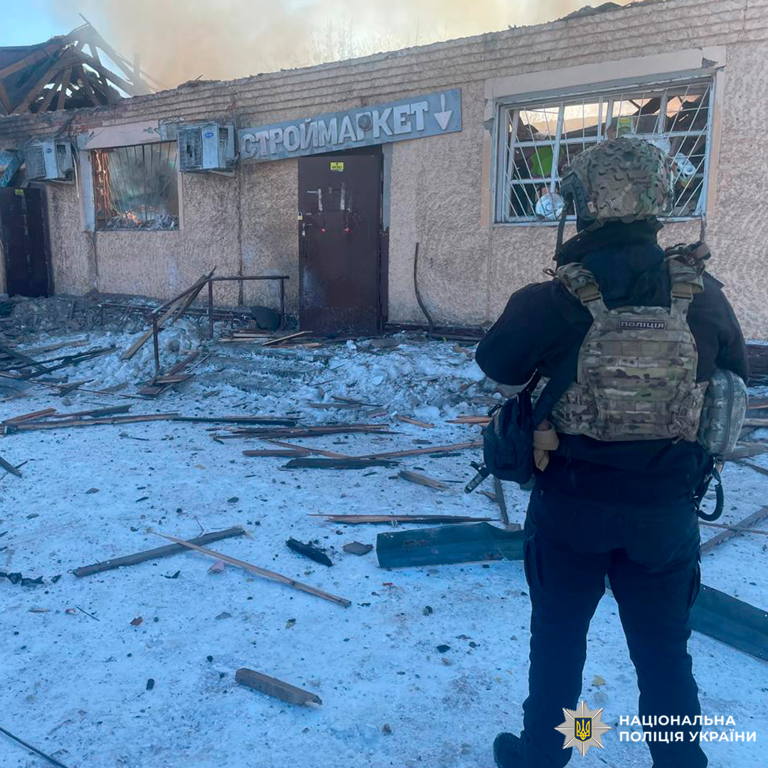 Поліцейський України оглядає зруйнований магазин «Строймаркет» із вибитими вікнами та уламками на засніженій території.