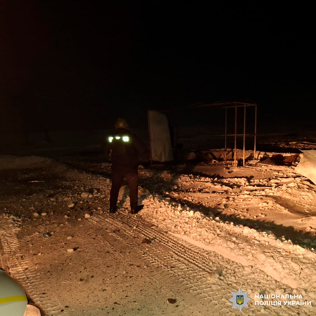 Поліцейський у світловідбивному спорядженні йде засніженою територією біля пошкодженої будівлі вночі.