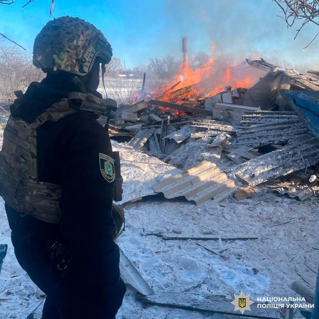Співробітник Національної поліції України у тактичному спорядженні, який контролює ситуацію біля зруйнованої та охопленої вогнем споруди.